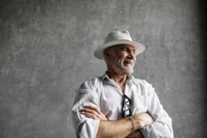 Elderly man smiling with folded arms, wearing a white fedora in a studio setting.